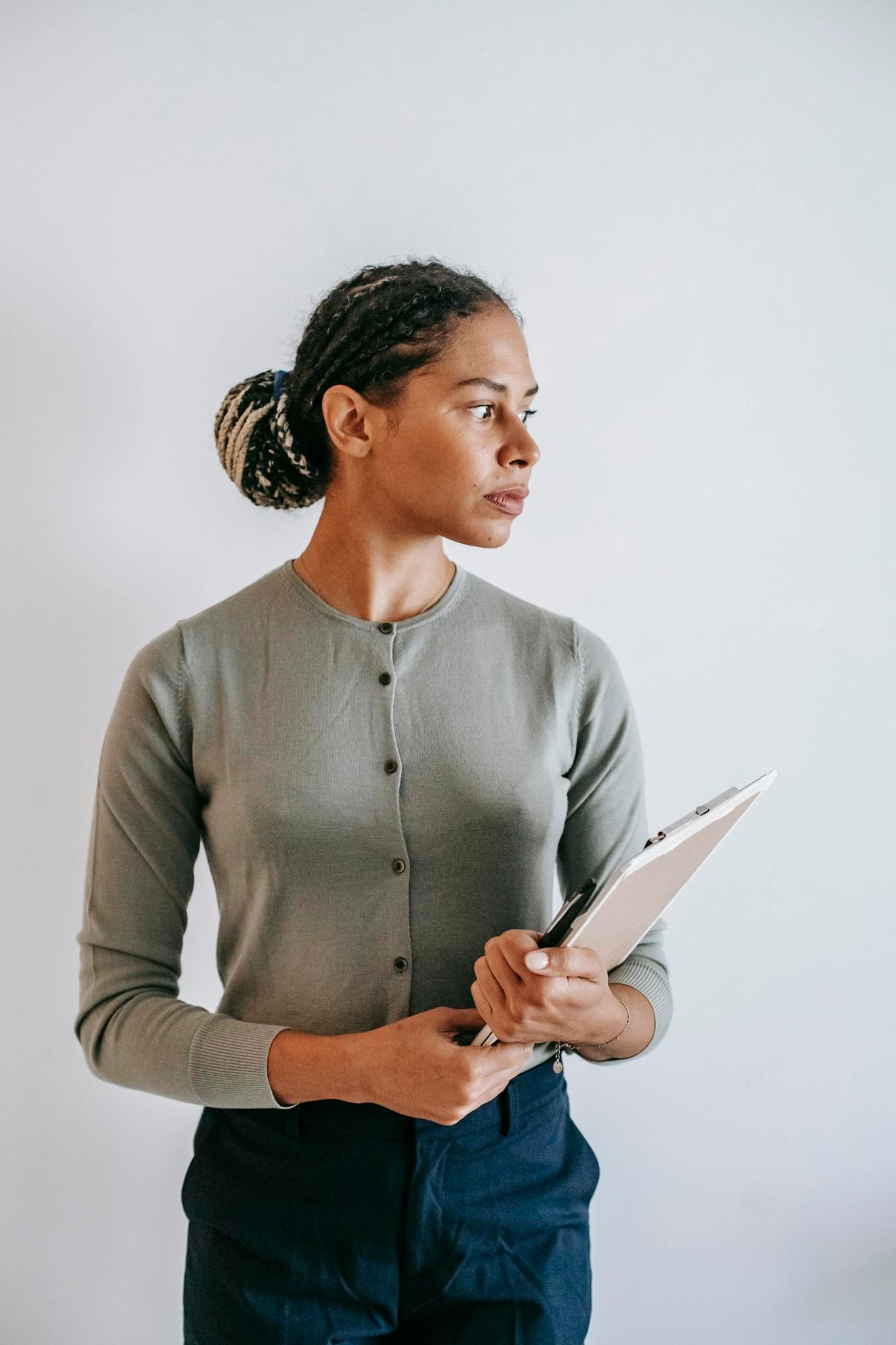 Serious ethnic female in formal wear with clipboard in hands standing against white wall and looking away in contemplation